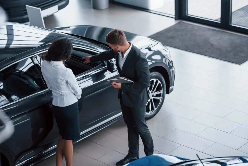 Salesperson showing a customer a car in a dealership showroom, representing dealer add-ons pitched in the finance office